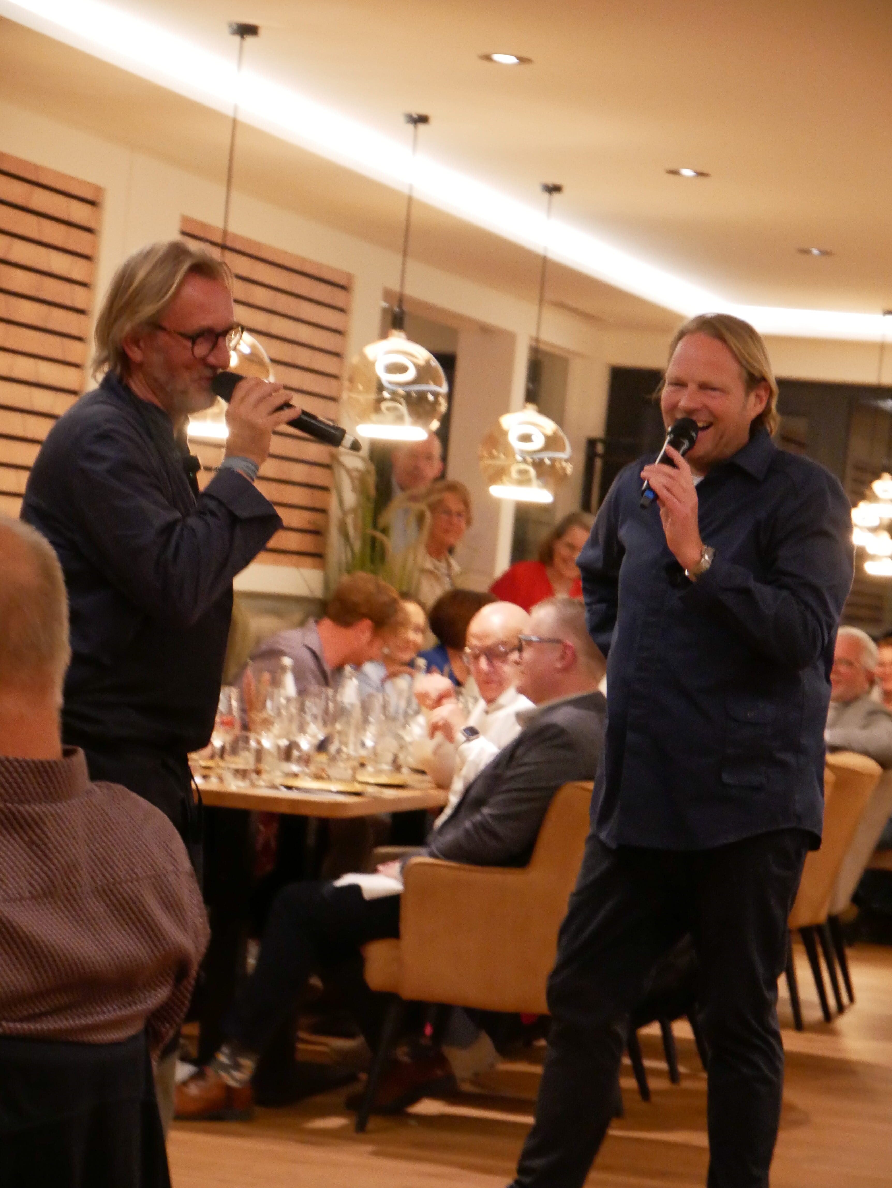 Zwei Männer singen in Mikrofone in einem Raum mit Menschen im Hintergrund.