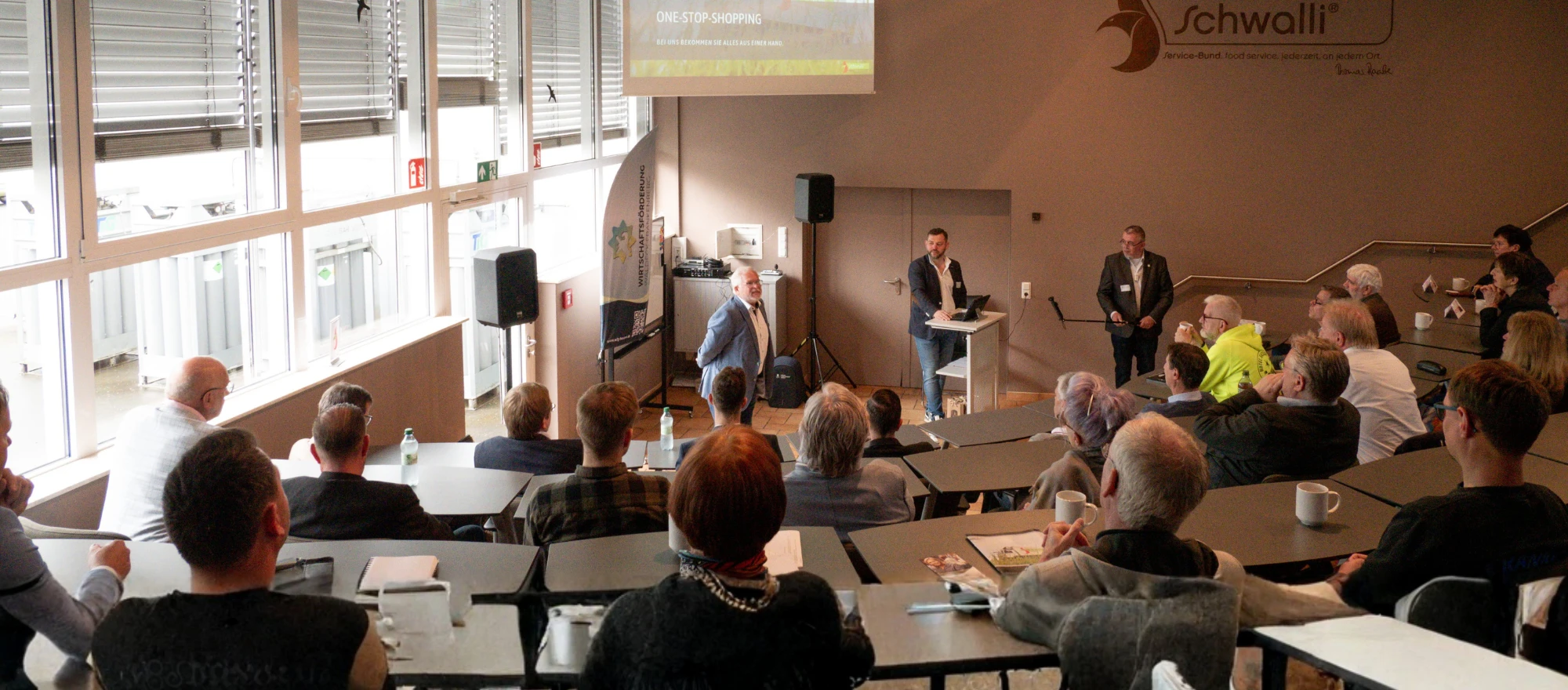Ein Mann steht vor einer Gruppe von Menschen in einem Raum mit einer Projektionsleinwand.