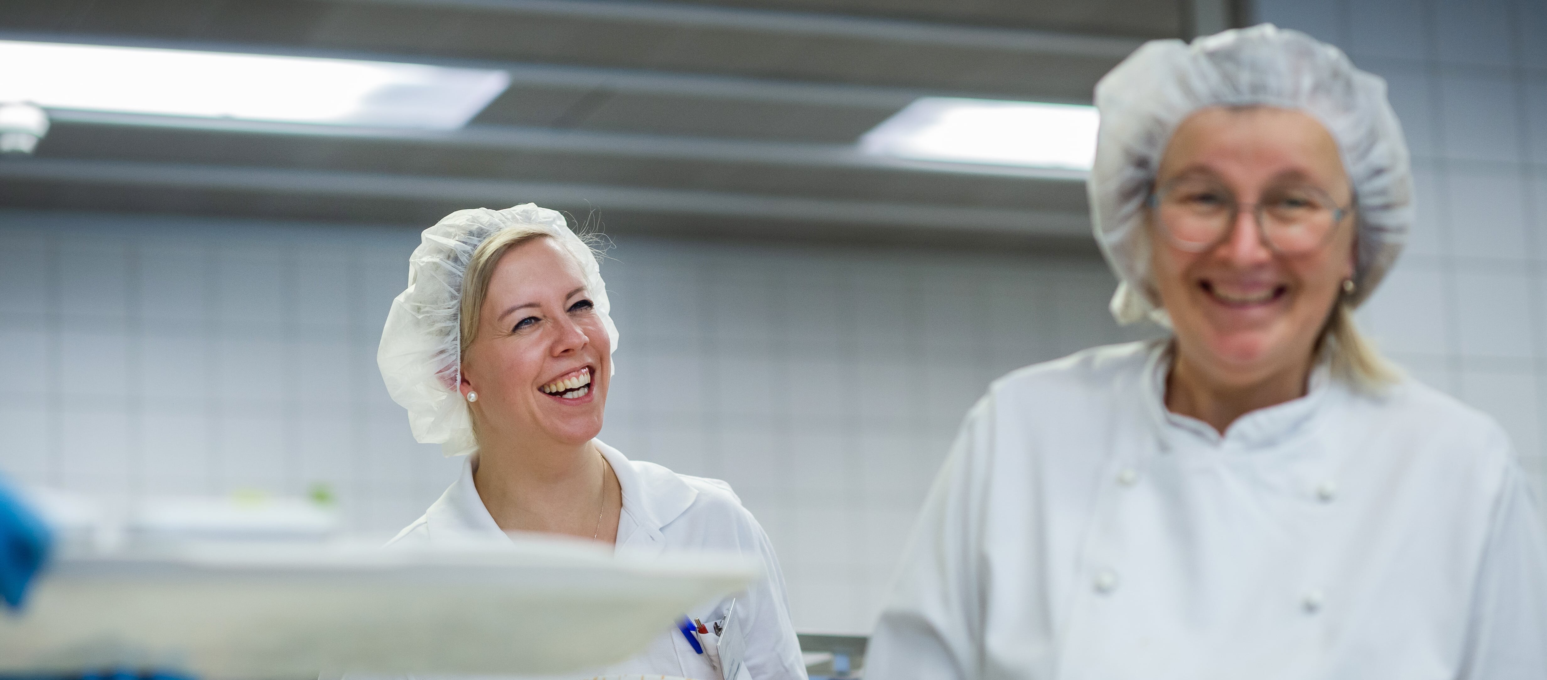 Zwei Frauen in weißen Kitteln und Haarnetzen lächeln sich an.