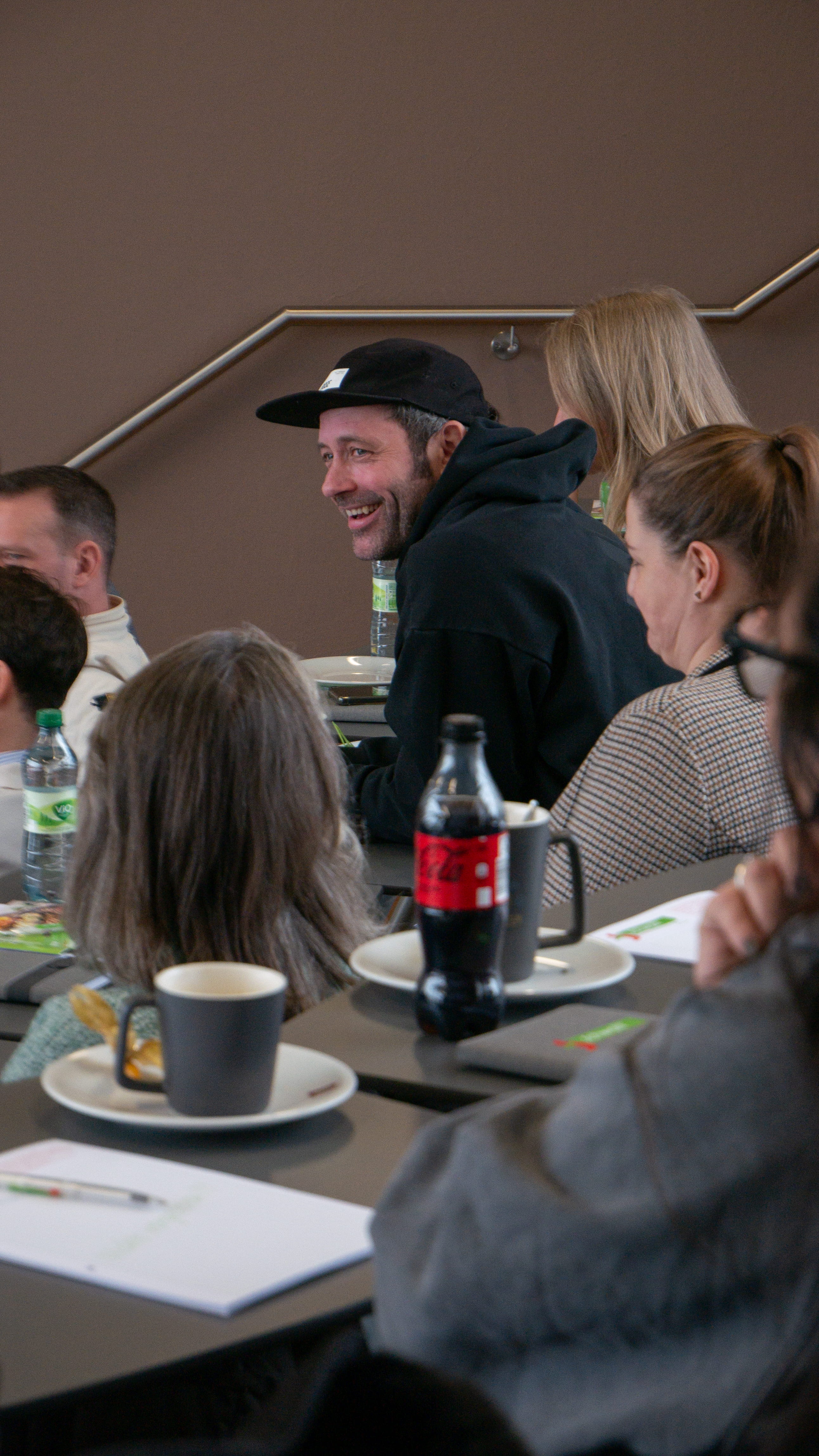 Erfolgsrezepte für Morgen Ein Mann mit schwarzem Hut lacht an einem Tisch mit mehreren sitzenden Personen.