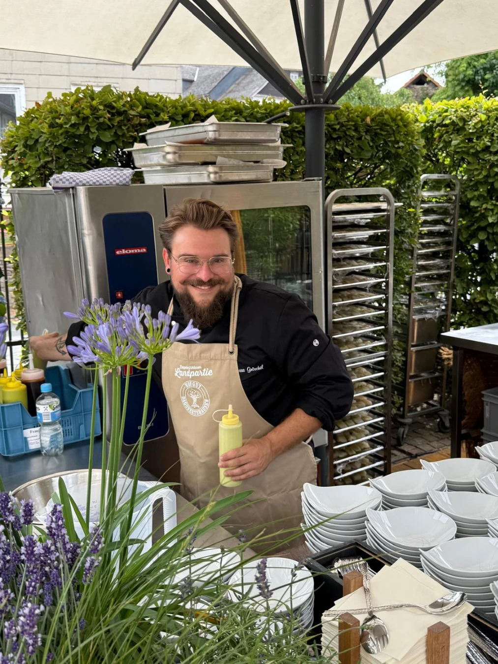 Tobias Gröschel Ein Mann in Schürze steht hinter einem Tisch mit Tellern und einer großen Gruppe von Pflanzen.