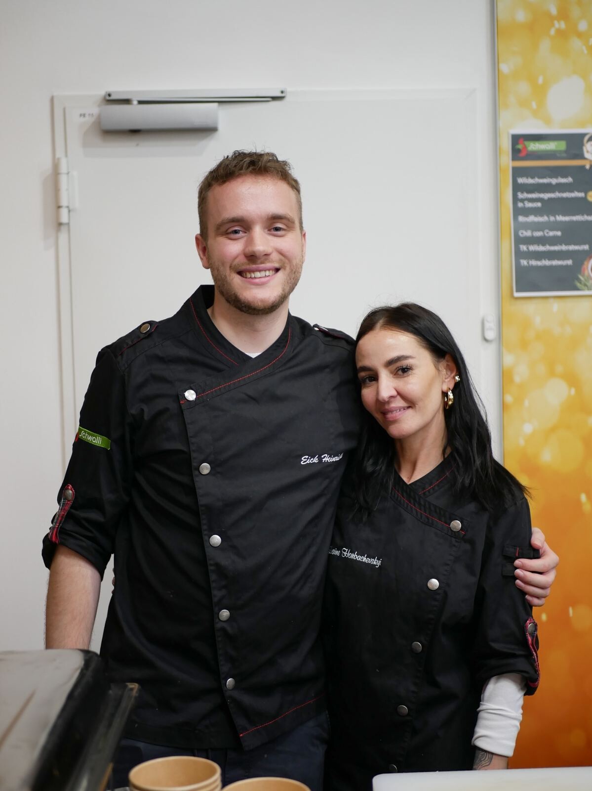 Ein Mann und eine Frau in schwarzen Uniformen lächeln in die Kamera.