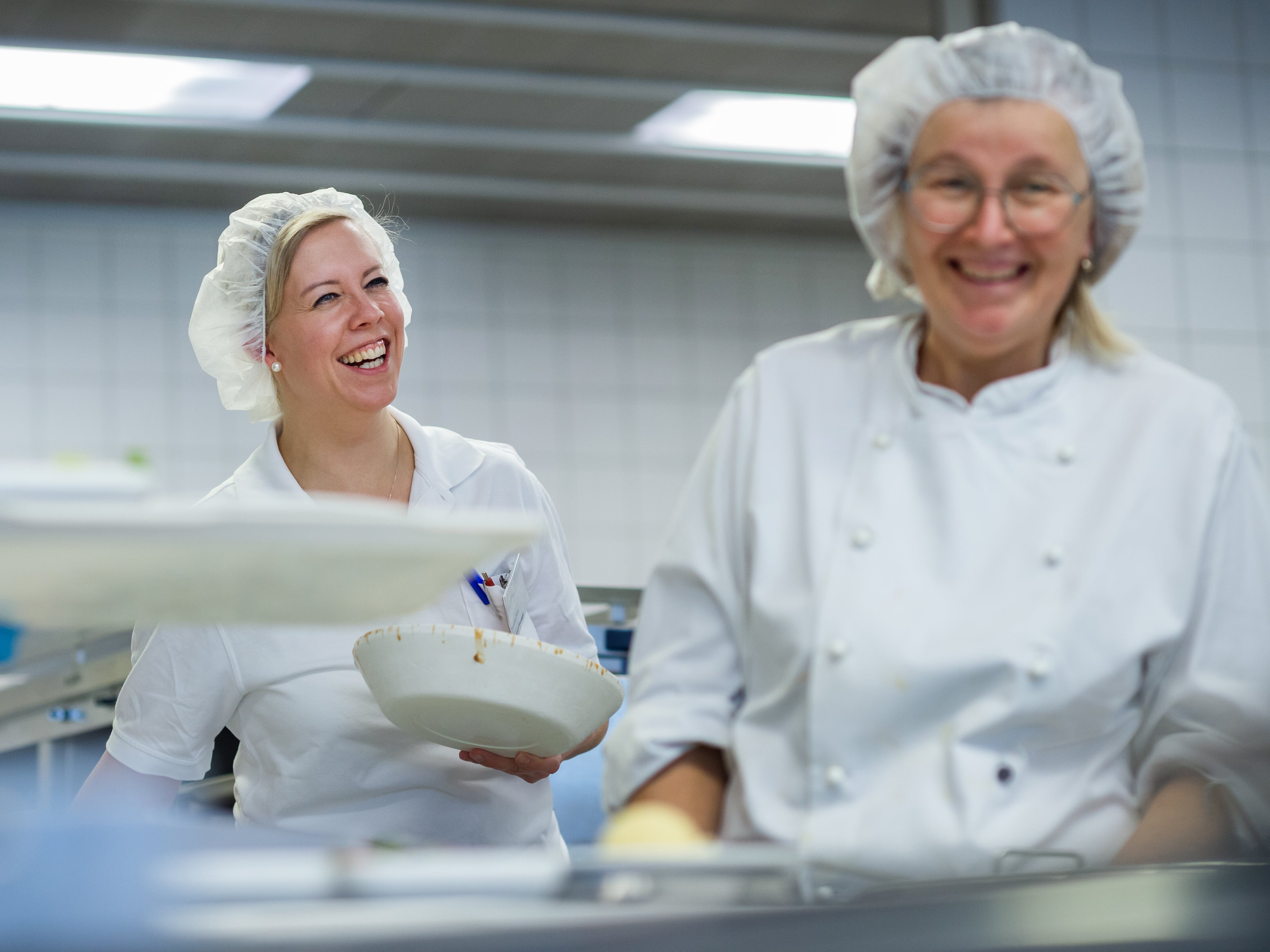 Stadtkrankenhaus Korbach Zwei Frauen in weißen Kitteln und Haarnetzen lächeln sich an.