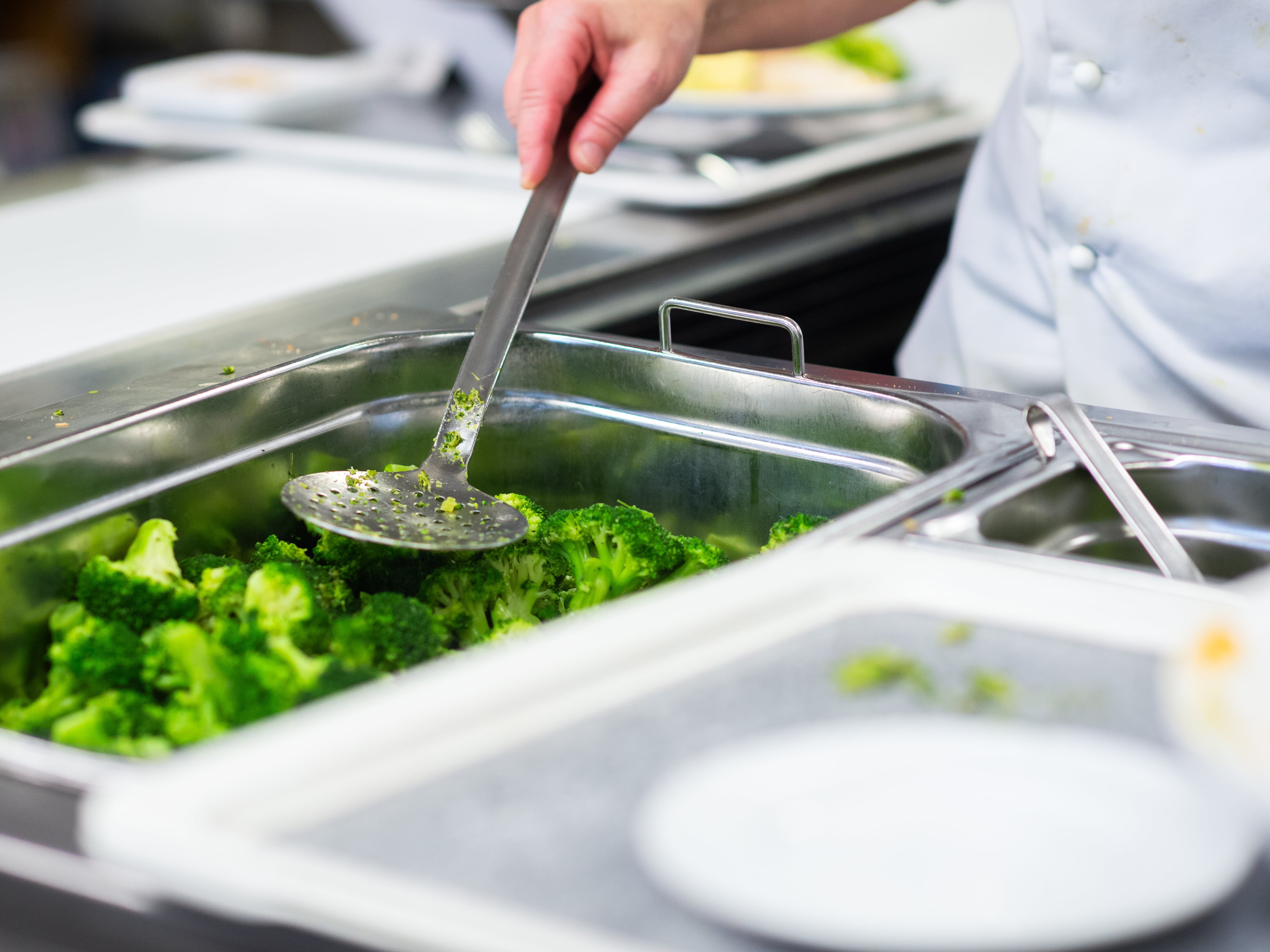 Eine Person hält eine Kelle über ein Tablett mit Brokkoli.