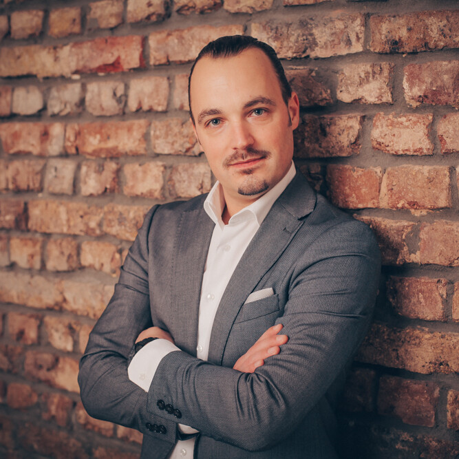 Markus Wessel Ein Mann lehnt lächelnd an einer Backsteinmauer und trägt formelle Kleidung.