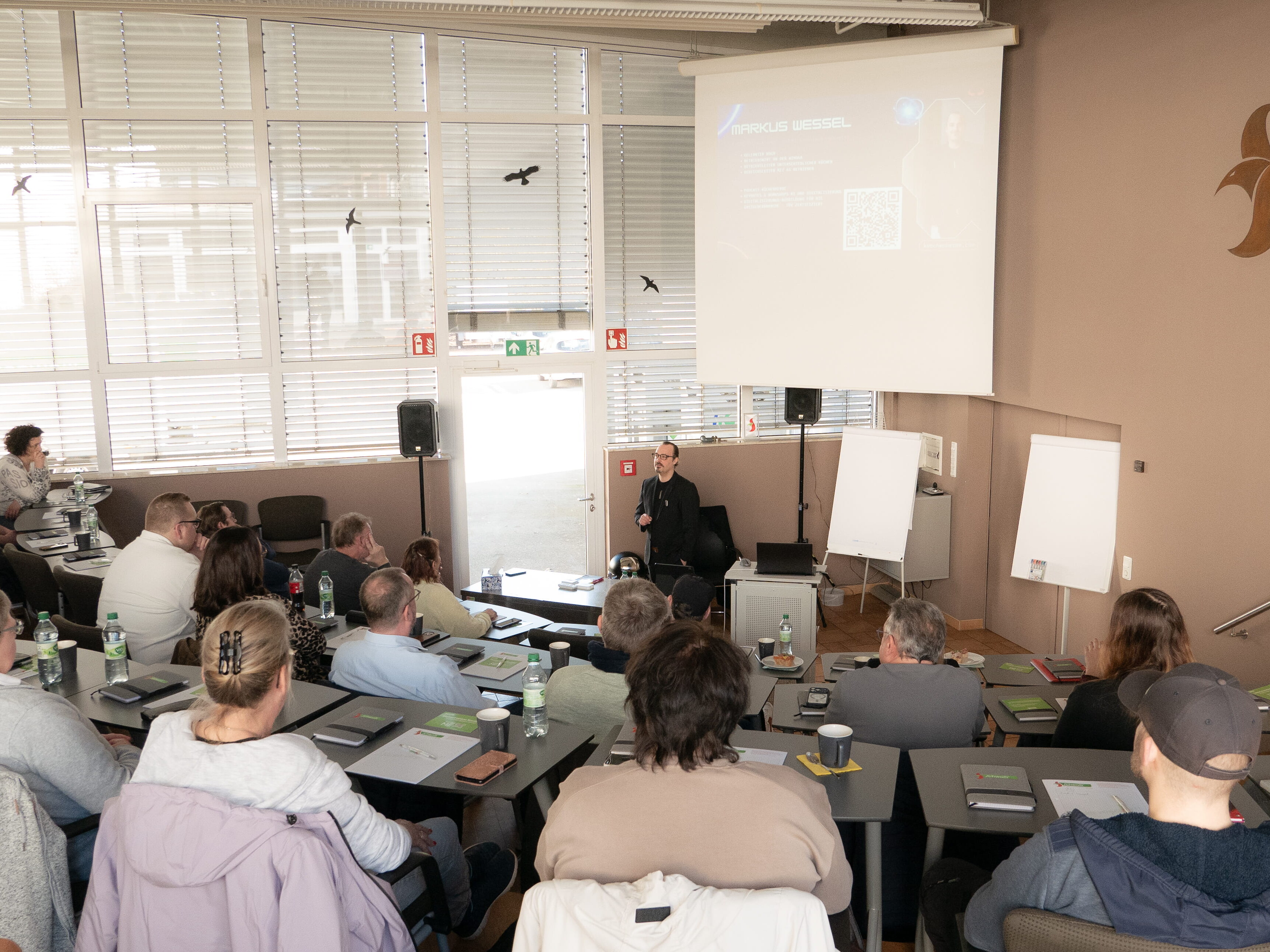 Ein Mann steht vor einer Gruppe von Menschen, die an Tischen sitzen.